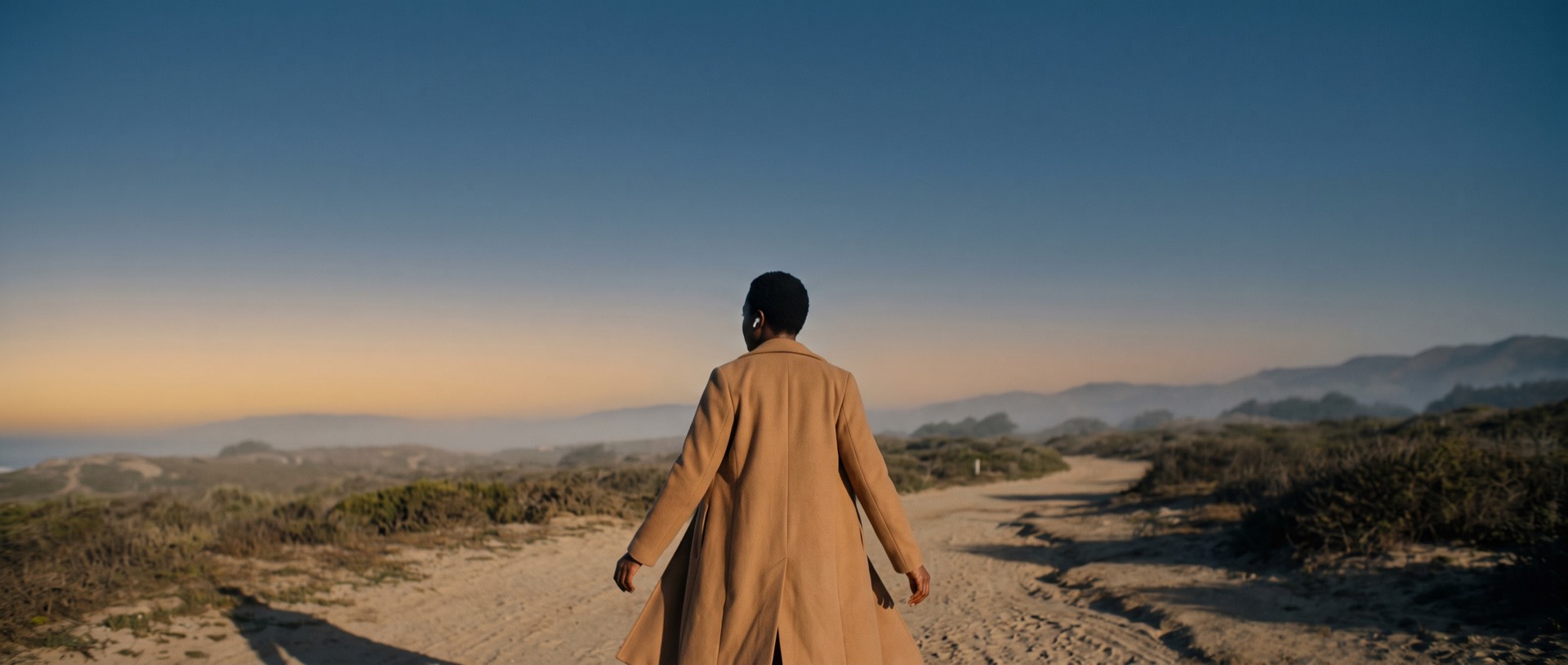 A woman in a long coat walks a coastal path into vast dawn light, speaking softly through an earbud.